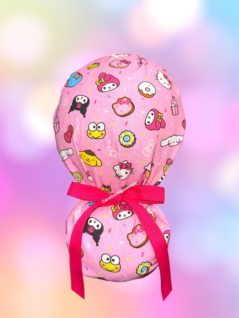 May include: A pink scrub cap featuring a repeating pattern of Sanrio characters including Hello Kitty, My Melody, Kuromi, Keroppi, and Pompompurin, interspersed with donuts and sprinkles. The cap is secured with a bright pink grosgrain ribbon.