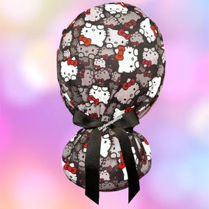 May include: A black and gray patterned scrub cap featuring white cartoon cat faces with red bows. The cap is tied with a black ribbon. The text "Carolboutiqueshops" is printed on the ribbon.