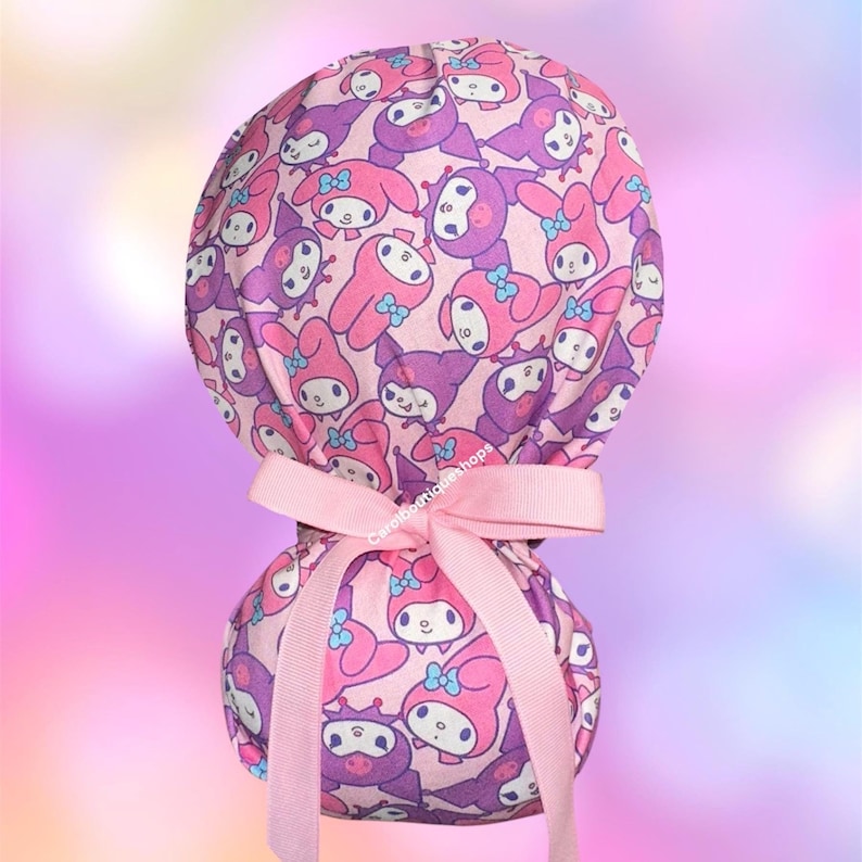 May include: A scrub cap featuring a repeating pattern of pink and purple cartoon characters with blue bows on a light pink background. The cap includes a light pink grosgrain ribbon tied in a bow at the back to secure the fit, with the text Carolboutiqueshops visible.