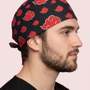 Puede incluir: Un gorro quirúrgico negro con un patrón repetitivo de nubes rojas con bordes blancos. El gorro presenta una forma redondeada e incluye cintas de tela en la parte posterior para ajustar el ajuste, proporcionando una cobertura completa de la cabeza.