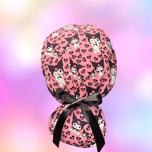 May include: A pink scrub cap featuring a repeating pattern of the character Kuromi and small black hearts. The cap has a gathered back and is secured with a black satin ribbon tied in a bow. The fabric displays a cartoon aesthetic throughout.