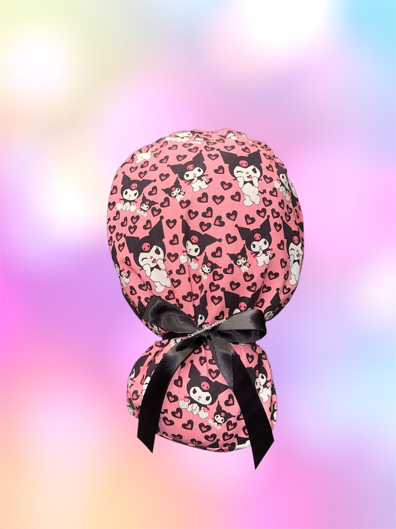 May include: A pink scrub cap featuring a repeating pattern of the character Kuromi and small black hearts. The cap has a gathered back and is secured with a black satin ribbon tied in a bow. The fabric displays a cartoon aesthetic throughout.