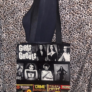 Puede incluir: Bolso tote negro con un collage de im&aacute;genes de pel&iacute;culas de terror vintage y el texto "Girls Will Be Ghouls". La bolsa presenta im&aacute;genes de personajes cl&aacute;sicos de pel&iacute;culas de terror y portadas de c&oacute;mics. La bolsa tiene correas negras.