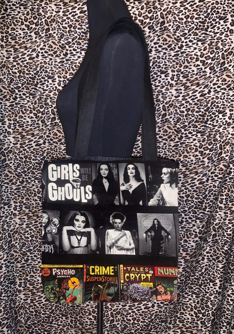 Puede incluir: Bolso tote negro con un collage de im&aacute;genes de pel&iacute;culas de terror vintage y el texto "Girls Will Be Ghouls". La bolsa presenta im&aacute;genes de personajes cl&aacute;sicos de pel&iacute;culas de terror y portadas de c&oacute;mics. La bolsa tiene correas negras.