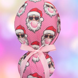 May include: A pink scrub cap featuring a repeating pattern of Santa Claus faces wearing heart-shaped sunglasses and blowing bubble gum. The cap is tied with a pink ribbon. The background is a soft, blurred gradient of colors.