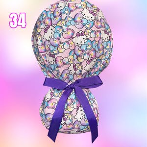 May include: A scrub cap featuring a repeating pattern of Hello Kitty characters and colorful bows on a light pink background. The cap is tied at the back with a solid purple grosgrain ribbon. The number 34 is printed in pink on the upper left.