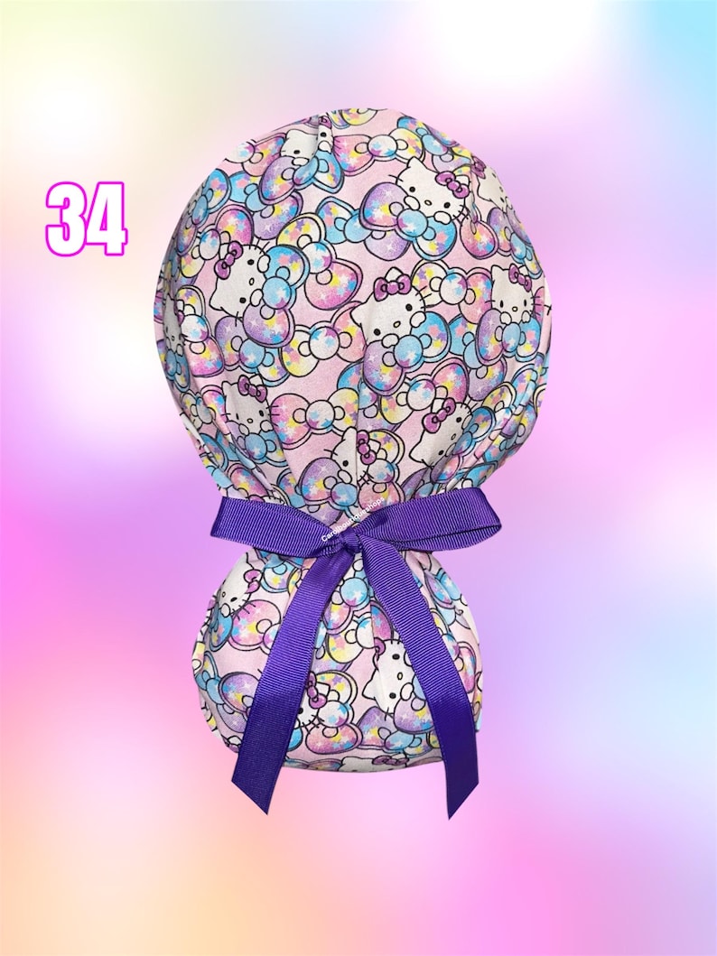 May include: A scrub cap featuring a repeating pattern of Hello Kitty characters and colorful bows on a light pink background. The cap is tied at the back with a solid purple grosgrain ribbon. The number 34 is printed in pink on the upper left.