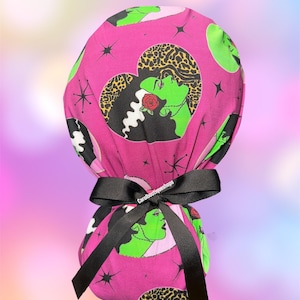 May include: A pink scrub cap featuring a repeating pattern of a green-skinned woman with black hair and a red rose, set against a black and leopard print heart. The cap is tied with a black ribbon. The text "Carolboutiqueshops" is printed on the ribbon.
