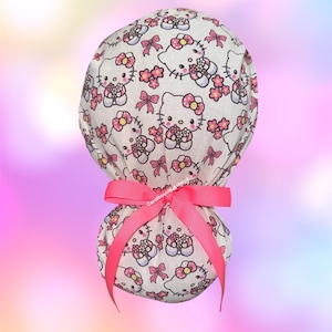 May include: A white scrub cap featuring a repeating pattern of Hello Kitty characters with pink bows and small pink flowers. The cap is gathered at the back and secured with a bright pink ribbon tied into a bow. The text Carolboutiqueshop is visible.