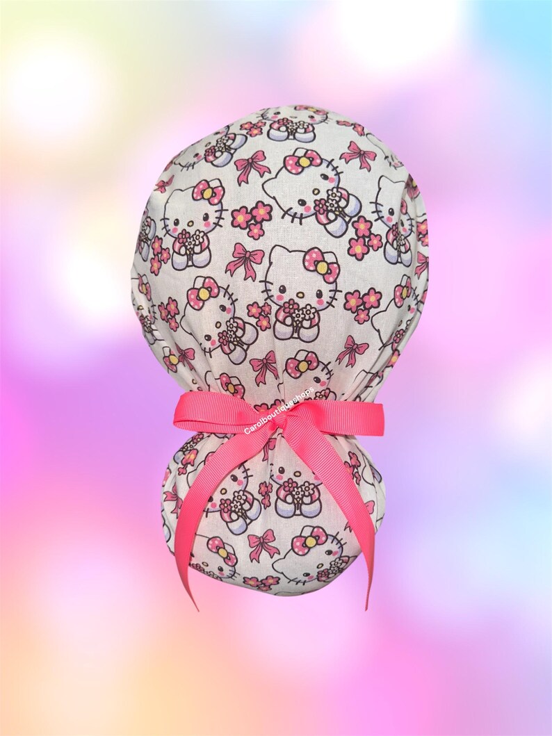 May include: A white scrub cap featuring a repeating pattern of Hello Kitty characters with pink bows and small pink flowers. The cap is gathered at the back and secured with a bright pink ribbon tied into a bow. The text Carolboutiqueshop is visible.