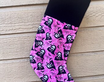 Puede incluir: Un calcetín de Halloween con puño negro y cuerpo rosa. El cuerpo del calcetín está estampado con la máscara de Ghostface de las películas Scream, bocadillos y murciélagos. El calcetín cuelga de un lazo negro.