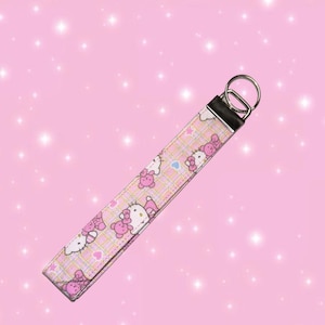 May include: A fabric wristlet keychain featuring a repeating pattern of Hello Kitty characters, pink bows, and small hearts on a light pink and yellow plaid background, finished with a silver-toned metal clasp and a circular split ring.