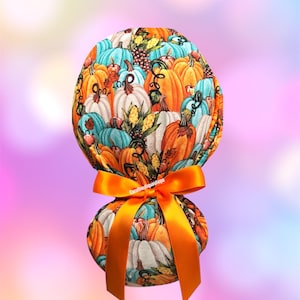 May include: A fabric ponytail holder featuring a colorful pumpkin pattern in shades of orange, teal, and white. The scrunchie is tied with a bright orange satin ribbon. The background is a soft gradient of pink, purple, and yellow.