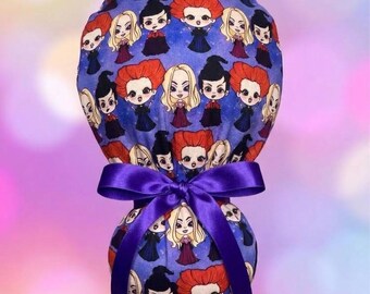 May include: A blue fabric scrub cap featuring a repeating pattern of cartoon characters with red, blonde, and black hair. The cap has a gathered back section secured with a vibrant purple satin ribbon tied into a bow at the center of the fabric.