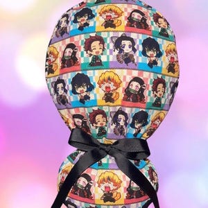 May include: A scrub cap featuring a colorful grid pattern with various anime character illustrations in shades of yellow, red, purple, and teal. The fabric head covering is gathered at the back and secured with a black ribbon tied in a bow.