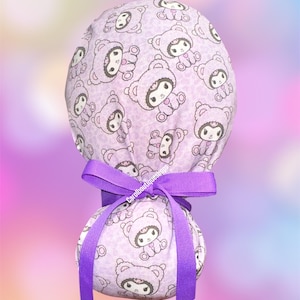 May include: A light purple scrub cap featuring a repeating pattern of cartoon characters wearing bear hoods. The cap is secured with a vibrant purple grosgrain ribbon tied in a bow at the back. The fabric has a subtle animal print background texture.