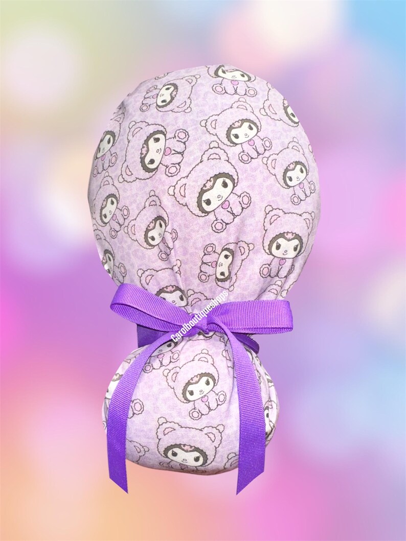 May include: A light purple scrub cap featuring a repeating pattern of cartoon characters wearing bear hoods. The cap is secured with a vibrant purple grosgrain ribbon tied in a bow at the back. The fabric has a subtle animal print background texture.