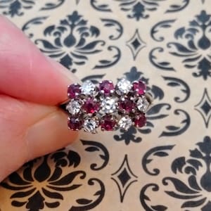 May include: A silver ring with a cluster of small, round gemstones. The ring features alternating red and clear stones, creating a sparkling, eye-catching design. The ring is held against a patterned background.