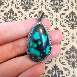 May include: A teardrop-shaped pendant featuring a polished dark stone with vibrant turquoise-colored inclusions. The stone is set in a silver-toned metal frame with a twisted rope border and decorative leaf-like accents at the base and a bail.
