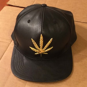 May include: Black baseball cap with a flat peak and a gold-coloured cannabis leaf emblem. The cap is made of a leather-like material and has a button on top. A fashionable headwear item.