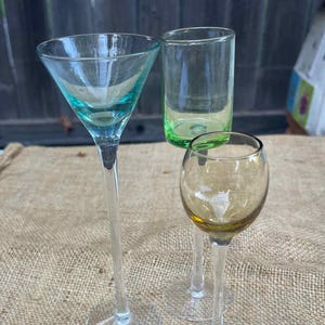 Może przedstawiać: Trzy różne kolorowe szklanki do picia. Wysoki kieliszek do martini z turkusową miseczką, krótka zielona szklanka do shotów i mały bursztynowy kieliszek do wina. Wszystkie szklanki mają przezroczyste nóżki i podstawy.