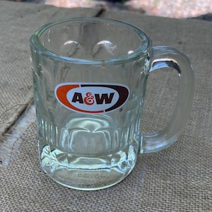 May include: A clear glass mug with a thick handle and a textured base featuring the A&W logo printed in orange, brown, and white on the front. The mug has a cylindrical shape with vertical indentations around the lower portion of the glass body.