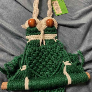 May include: Green and white macrame wall hanging with wooden beads and a wooden dowel. The hanging has a fringe at the bottom and is labeled "Frog Plant Hanger".