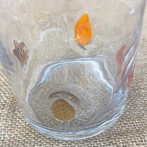 May include: Clear glass tumbler with embossed autumn leaf designs in shades of orange, brown, and gold. The glass has a textured surface and is set against a neutral background.