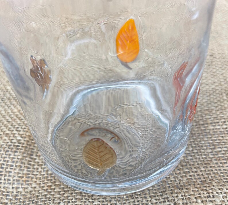 May include: Clear glass tumbler with embossed autumn leaf designs in shades of orange, brown, and gold. The glass has a textured surface and is set against a neutral background.