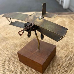 Op de afbeelding: Een klein, handgemaakt metalen vliegtuigbeeldje met een houten voet. Het vliegtuig is gemaakt van messing en heeft een propeller en wielen. De voet is van donkerbruin hout.