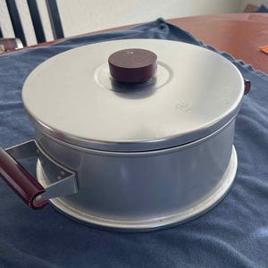 May include: A silver aluminum cake pan with a removable bottom and a maroon handle. The pan has a maroon knob on the lid.