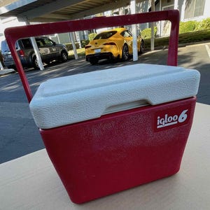 Puede incluir: Una nevera Igloo roja y blanca con asa de transporte. La nevera tiene una tapa blanca y una base roja con el logotipo de Igloo y el número 6 impreso en ella.