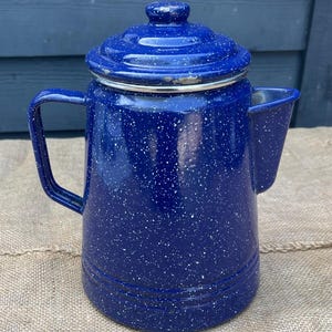 May include: A blue speckled enamelware coffee pot with a lid and handle. The pot has a spout for pouring and a silver rim. The pot is sitting on a burlap surface.