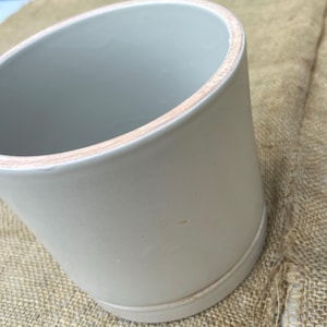 Puede incluir: Una jarra de cerámica gris claro con una forma ligeramente acampanada. El borde tiene un borde natural sin esmaltar. La jarra se asienta sobre una base con una sutil cresta. El fondo es una tela texturizada de color beige.