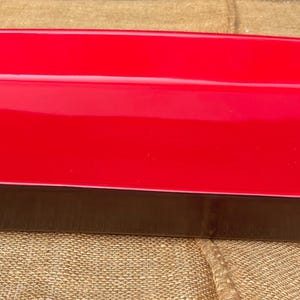 May include: A rectangular serving tray with a glossy red interior and a dark brown exterior. The tray is empty and sits on a textured, tan surface.