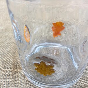 May include: Clear glass tumbler with embossed orange and gold autumn leaves. The glass has a textured surface and is partially filled with liquid. The leaves are in various shapes and sizes, adding a seasonal touch to the glassware.