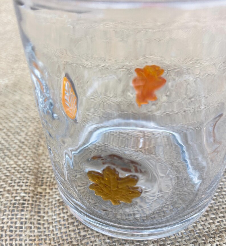 May include: Clear glass tumbler with embossed orange and gold autumn leaves. The glass has a textured surface and is partially filled with liquid. The leaves are in various shapes and sizes, adding a seasonal touch to the glassware.