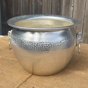May include: A silver metal ice bucket with a hammered texture around the middle. The bucket has two lion head handles with rings. The bucket is sitting on a brown burlap surface.