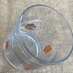 May include: A clear glass tumbler with orange and brown floral designs. The glass has a cylindrical shape and is slightly tilted, showcasing the interior. The floral designs are evenly spaced around the glass.