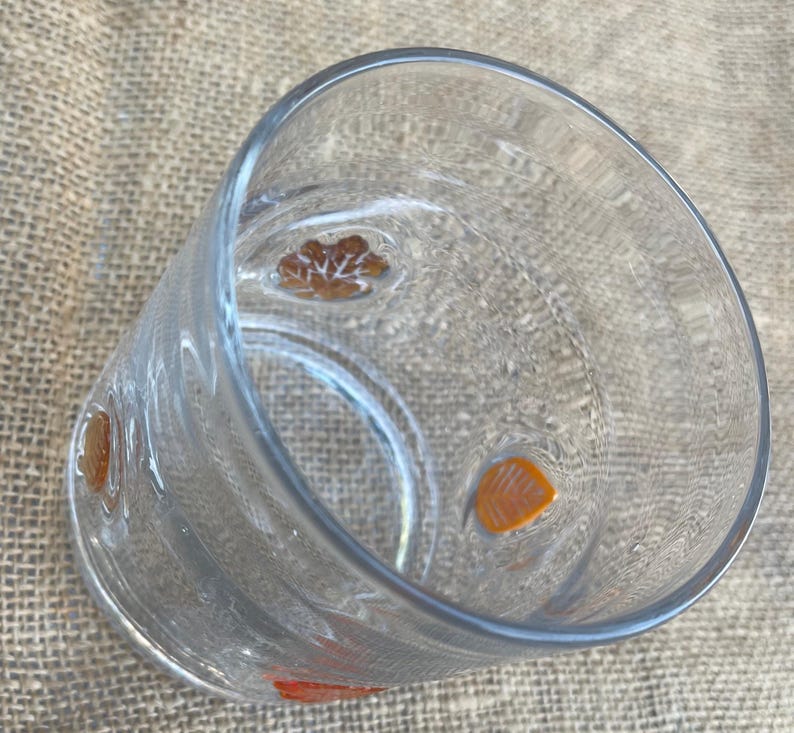 May include: A clear glass tumbler with orange and brown floral designs. The glass has a cylindrical shape and is slightly tilted, showcasing the interior. The floral designs are evenly spaced around the glass.