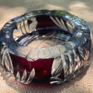 May include: A round, heavy glass ashtray featuring a deep ruby red center and a thick, clear glass rim. The exterior of the rim is decorated with a series of vertical, carved, leaf-like patterns that catch the light, creating a textured appearance.