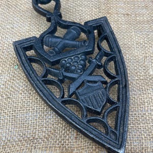 May include: Black cast iron trivet with a handle. The trivet features a shield, crossed swords, grapes, and bottles design. The trivet is shaped like an iron and has an openwork design.