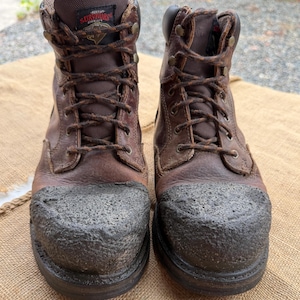 Puede incluir: Un par de botas de trabajo de cuero marrón con punteras reforzadas de textura oscura, cordones marrones y una etiqueta negra en la lengüeta con el texto Herman Survivors. Las botas tienen un diseño de caña alta con ojales metálicos y construcción.
