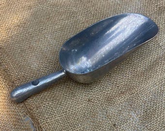 Può includere: Una paletta in metallo argentato con una ciotola arrotondata e un manico con un foro. La paletta è realizzata in metallo lucido e riflettente ed è posta su uno sfondo beige strutturato.