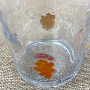 May include: A clear glass tumbler with a textured surface. The glass is decorated with several orange and gold leaf designs. The leaves are in various shapes and sizes, adding a seasonal touch to the glassware.