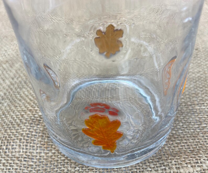 May include: A clear glass tumbler with a textured surface. The glass is decorated with several orange and gold leaf designs. The leaves are in various shapes and sizes, adding a seasonal touch to the glassware.