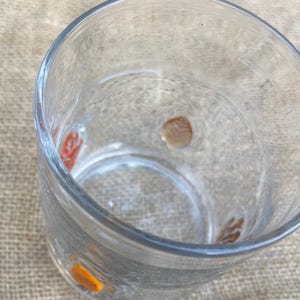 May include: A clear glass with decorative orange, red, and brown accents. The glass is empty and sits on a neutral-colored surface. The glass has a thick base and a wide opening.