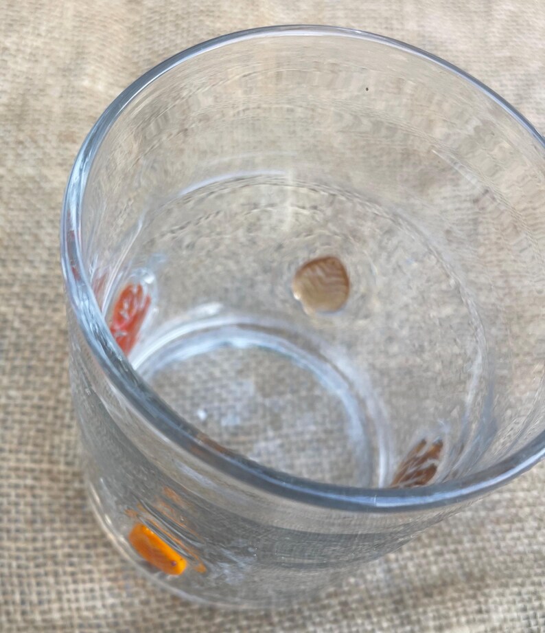 May include: A clear glass with decorative orange, red, and brown accents. The glass is empty and sits on a neutral-colored surface. The glass has a thick base and a wide opening.