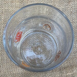 May include: Clear glass tumbler with embossed floral designs in orange and red. The glass has a thick base and a slightly textured surface. The tumbler is empty and sits on a textured, tan surface.