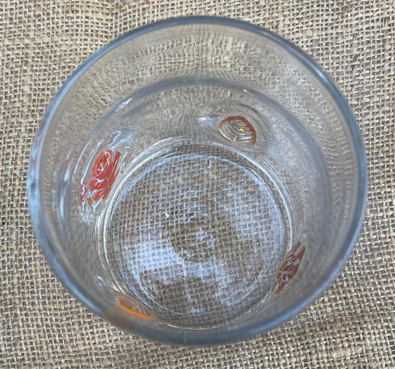 May include: Clear glass tumbler with embossed floral designs in orange and red. The glass has a thick base and a slightly textured surface. The tumbler is empty and sits on a textured, tan surface.